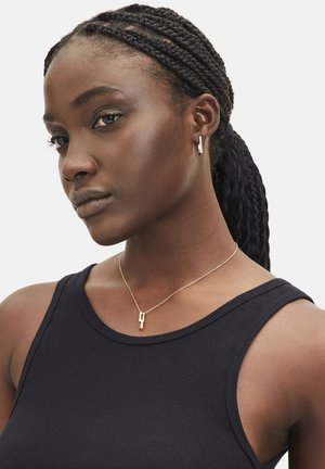 Gouden ketting met een geometrische hanger, gedragen over een zwart geribbelde tanktop. Vlecht haar en kleine gouden oorringen completeren de look.