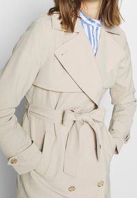 Béžový trenčkot s dvouřadým zap�ínáním, širokým límcem a páskem v pase. Má zlaté knoflíky a dvě boční kapsy.