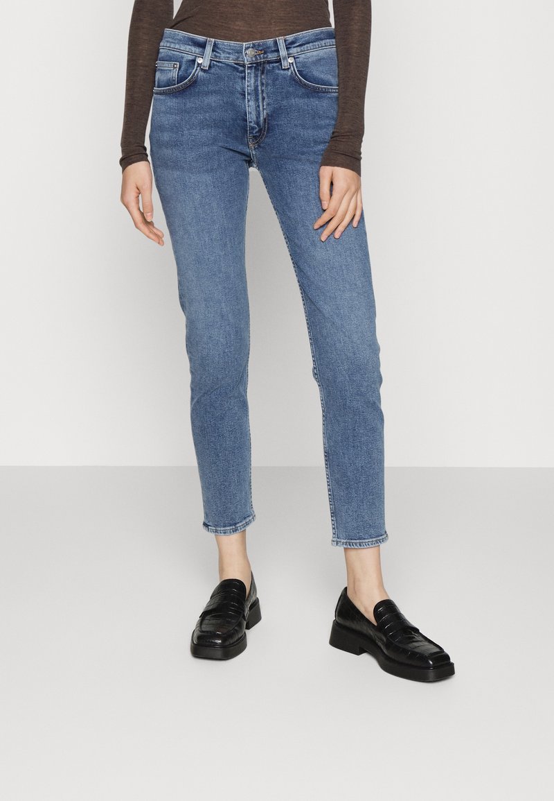 Modré skinny džíny s vysokým pasem, které mají pět kapes, minimální oděrky a rovný zkrácený lem. Kombinováno s černými mokasínami.