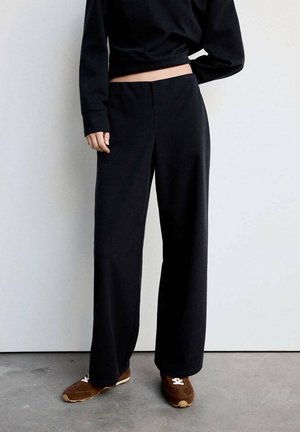 Persona con pantalones negros holgados y zapatillas marrones de pie contra una pared blanca sobre un suelo gris.