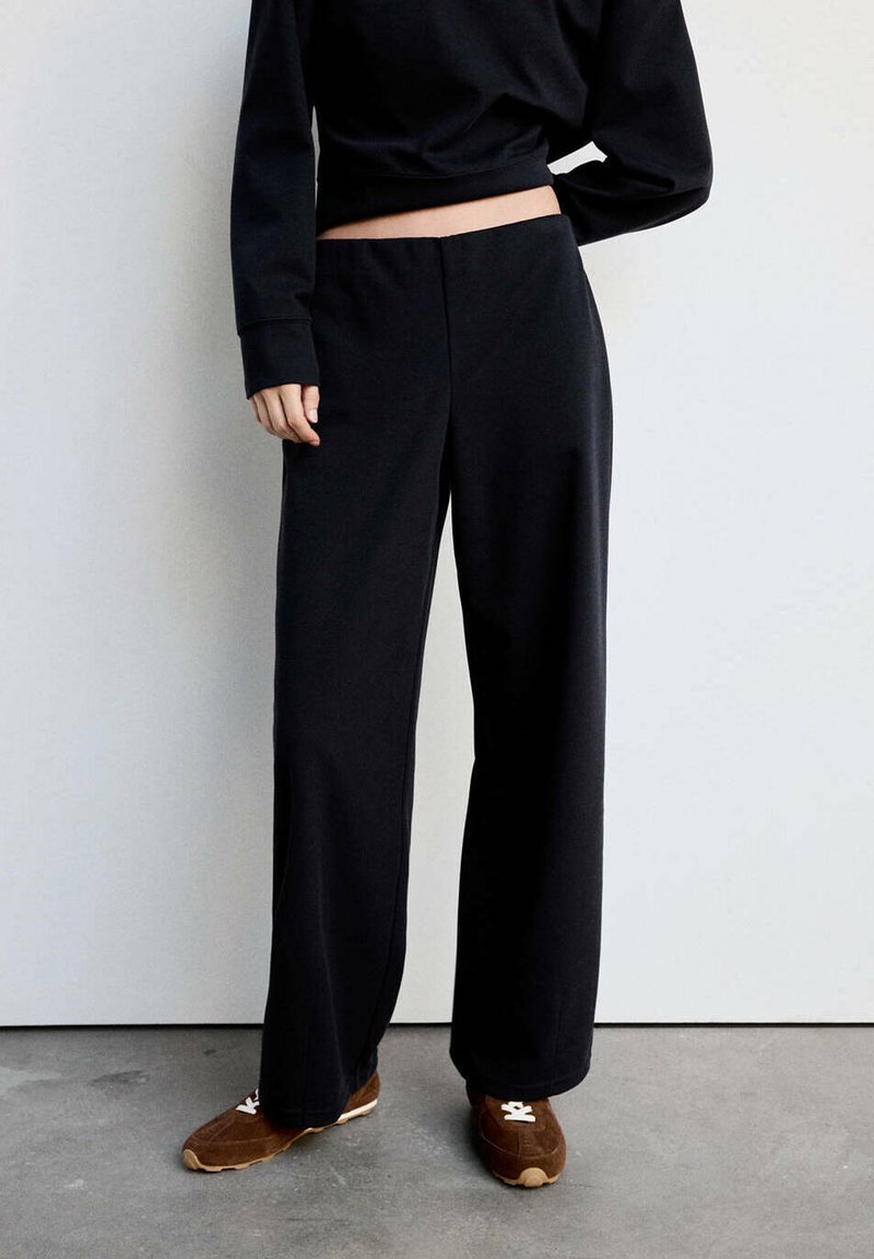 Personne portant un pantalon noir ample et des baskets marron, debout contre un mur blanc sur un sol gris.