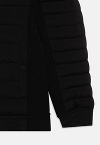 Section de veste noire matelassée avec un côté en tissu noir lisse. Comprend une poche à bouton et des poignets côtelés. Design texturé et élégant.