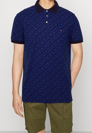 Polo blu navy con motivo a fantasia, abbottonatura a tre bottoni e colletto scuro; indossata con pantaloncini cargo verde oliva.