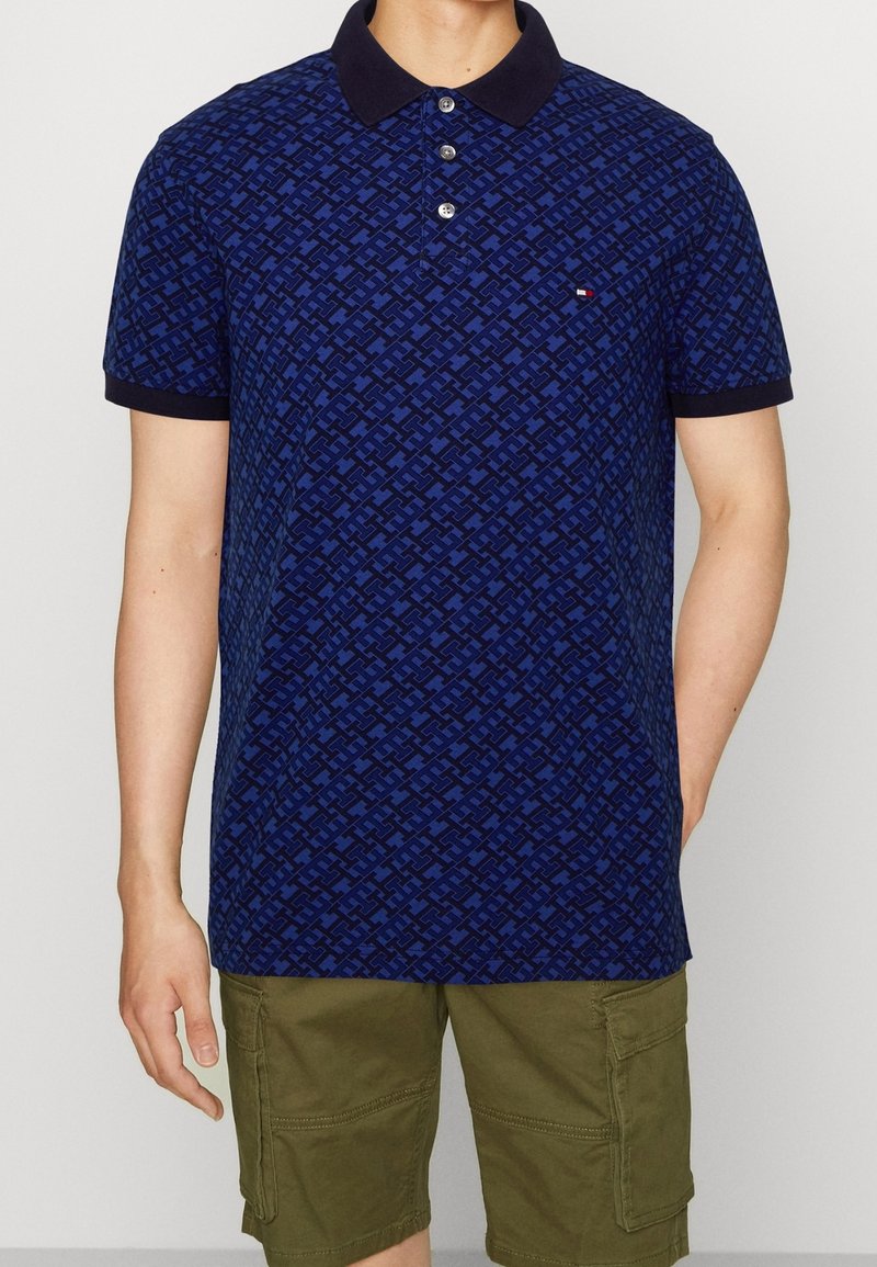 Polo bleu marine à motif, avec patte de boutonnage à trois boutons et col foncé ; porté avec un short cargo vert olive.