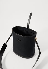 Bolso tipo cubo de lona negra con base de piel suave, una correa para el hombro resistente y un acabado en tono dorado. La parte superior abierta revela un interior espacioso.