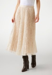 Jupe midi plissée en dentelle crème avec une finition texturée, dotée d'une taille et d'un détail de bouton. Associée à des bottes à talons brunes.