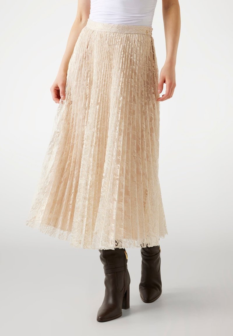 Jupe midi plissée en dentelle crème avec une finition texturée, dotée d'une taille et d'un détail de bouton. Associée à des bottes à talons brunes.