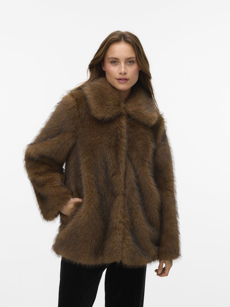 Moteris, vilkinti rudą dirbtinę kailinę apsiaustą su plačiomis apykaklėmis, viena ranka kišenėje, ant vientisos baltos fono.