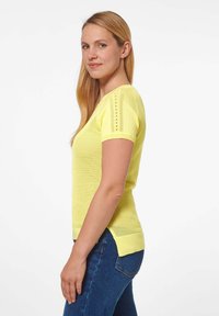 Geel gebreid T-shirt met korte mouwen, voorzien van een textuur patroon en zijsplitten. G gedragen over een paar donkere spijkerbroeken, wat een strakke pasvorm laat zien.