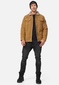 Bruine corduroy jas met een harige kraag, voorzien van twee borstzakken, gedragen over een zwarte trui en gecombineerd met donkere jeans en zwarte laarzen.