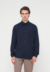 Σκούρο μπλε πουκάμισο με μακριά μανίκια, γιακά με κουμπιά και μαύρα κουμπιά, με λεία υφή. Συνδυάζεται με ανοιχτό καφέ παντελόνι.