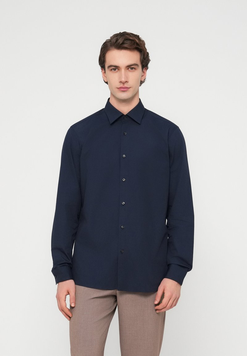 Σκούρο μπλε πουκάμισο με μακριά μανίκια, γιακά με κουμπιά και μαύρα κουμπιά, με λεία υφή. Συνδυάζεται με ανοιχτό καφέ παντελόνι.