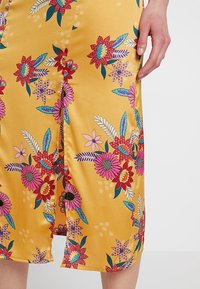 Gelbes Blumenkleid mit lebhaften Mustern aus blauen, pinken und roten Blumen, mit glänzender Textur und einem Schlitzdetail vorne.
