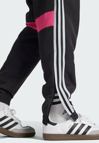 Schwarze Sporthosen mit weißen Seitestreifen und rosa Akzenten, kombiniert mit weißen Sneakers, die schwarze Streifen und braune Sohlen haben.