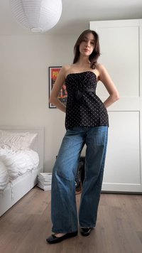 Kvinna står i ett sovrum iförd en axelbandslös svart topp med prickar, vida blå jeans och svarta ballerinaskor, med händerna på höfterna.