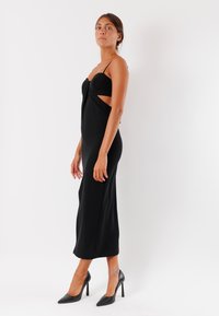 Abito aderente nero con design cut-out e spalline sottili. Tessuto liscio, lungo fino al ginocchio e senza motivi visibili. Indossato con tacchi alti neri.