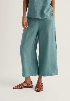 Persoon draagt losse teal cropped broek en bruine sandalen met bandjes en platte zool, staat met één hand gedeeltelijk in de zak.