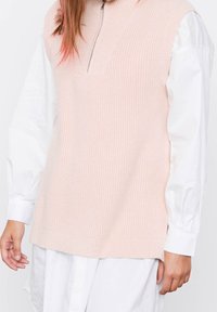 Kurzes pinkfarbenes ärmelloses Strickoberteil mit hohem Kragen und halbem Reißverschluss, über einem weißen Langarmshirt getragen, das strukturierter Stoff aufweist.