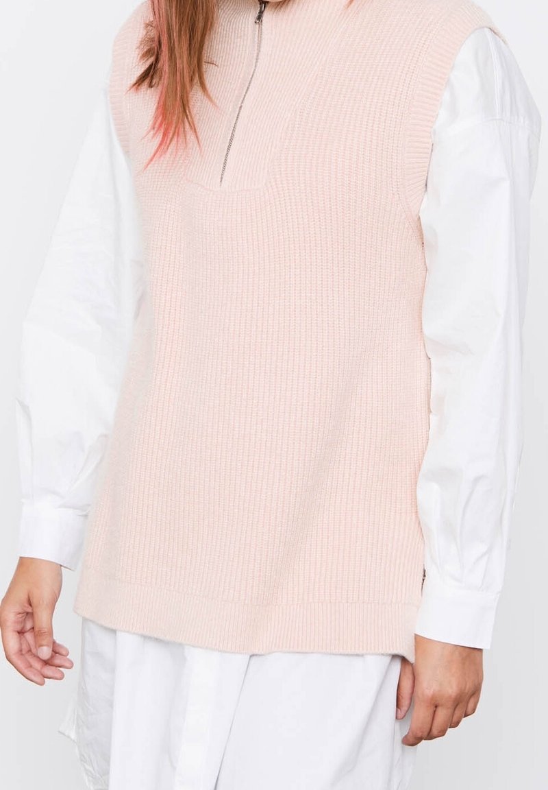 Kurzes pinkfarbenes ärmelloses Strickoberteil mit hohem Kragen und halbem Reißverschluss, über einem weißen Langarmshirt getragen, das strukturierter Stoff aufweist.