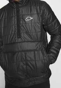 Zwarte gevoerde jas met een hoge kraag, met een kwart rits en een wit Nike-logo op de borst. Textuur met een doorgestikte ontwerp.