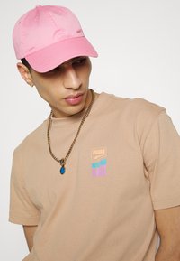 Casquette en coton rose clair avec sangle ajustable. T-shirt beige avec un logo graphique violet et bleu, associé à un collier en or et un pendentif.