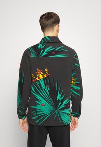 Czarna polarowa koszula z zielonymi wzorami liści palmowych, czerwonymi kropkami i żółtą koroną. Posiada kołnierzyk i mankiety zapinane na guziki.