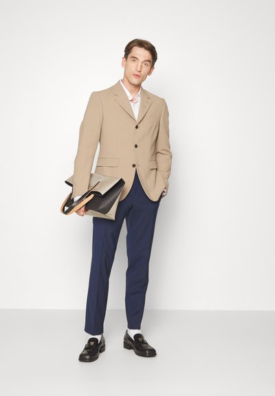 Blazer bege texturizado com botões pretos, calças azul-marinho e mocassim preto. Segurando uma bolsa bicolor com acabamentos em couro.