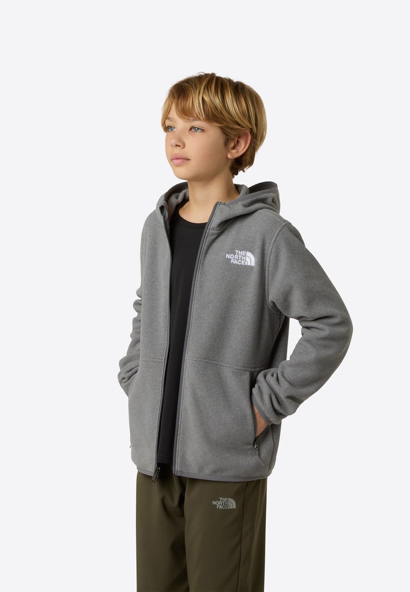 Sivá fleecová mikina na zips s kapucňou, prednými vreckami a vyšívaným logom na hrudi. Nosená cez čierne tričko a skombinovaná s olivovo zelenými nohavicami.