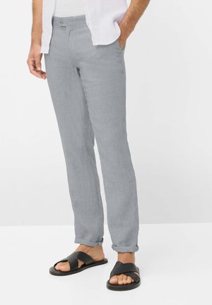 Man draagt lichtgrijze linnen broek met opgerolde pijpen en zwarte sandalen met kruisbandjes, staand met één hand in de broekzak.