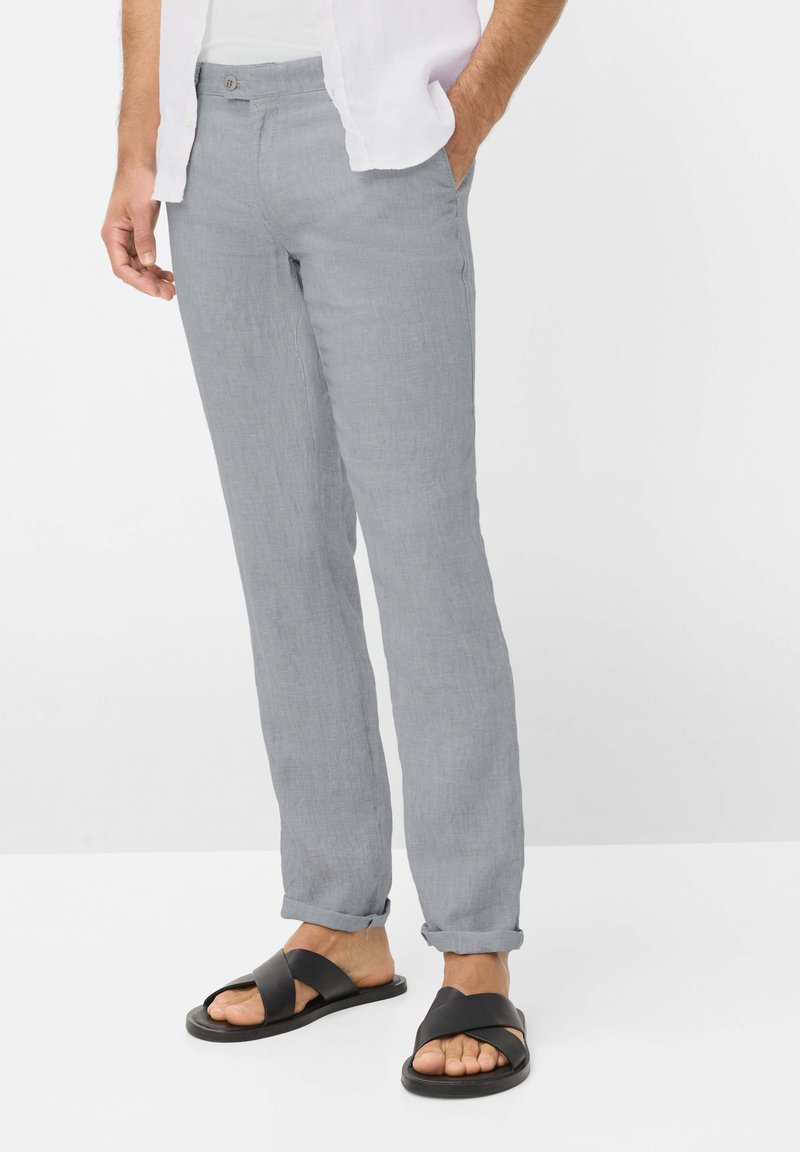 Mann trägt hellgraue Leinenhose mit hochgekrempelten Säumen und schwarzen Sandalen mit Kreuzriemen, steht mit einer Hand in der Hosentasche.