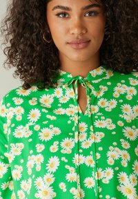 Blusa verde con un motivo floreale di margherite bianche con centri gialli. Legatura al collo e apertura a chiave nella parte anteriore.