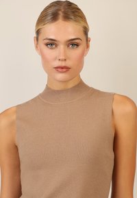 Beige gestricktes, ärmelloses Oberteil mit hohem Stehkragen. Glatte Textur und figurbetontes Design, mit subtilen horizontalen Linien über den Stoff.