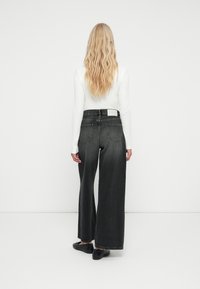 Široke črne jeans hlače z visokim pasom, ki imajo dve zadnji žepi in belo oznako blagovne znamke na hrbtnem pasu.