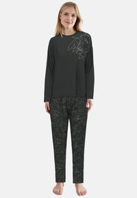 Young woman standing barefoot wearing dark long-sleeve top with floral outline on shoulder and matching floral patterned pants.