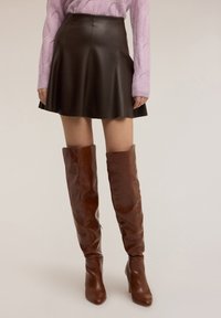 Brązowa skórzana spódnica w kształcie litery A, z teksturowanej tkaniny, w zestawieniu z brązowymi kozakami za kolano i jasnofioletowym swetrem z grubym splotem.