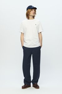White cotton t-shirt with a small logo, paired with loose navy chinos and brown moccasin shoes, worn with a dark blue cap.