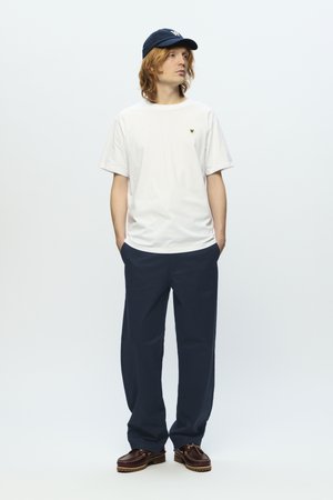 White cotton t-shirt with a small logo, paired with loose navy chinos and brown moccasin shoes, worn with a dark blue cap.