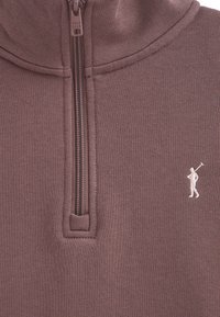 Sudadera de cremallera en color púrpura claro con cuello alto, que presenta un pequeño logo blanco de golfista y una tela texturizada. Incluye detalles de cremallera.