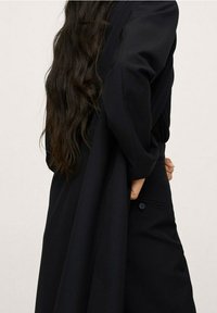 Vrouw met lang golvend donker haar, gekleed in een losse zwarte jas, terwijl ze de jas achter haar rug vasthoudt tegen een effen lichte achtergrond.