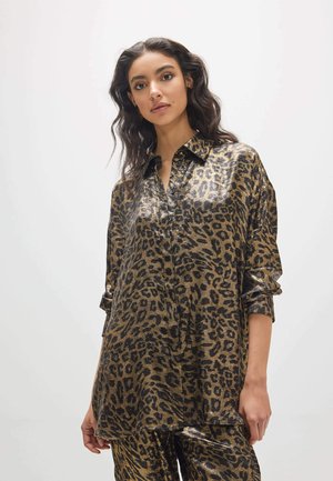 Mujer con cabello oscuro y ondulado que lleva una camisa de manga larga con estampado de leopardo en negro y dorado brillante y pantalones a juego sobre un fondo liso.