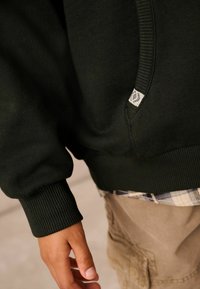 Tmavo zelená mikina so žebrovanými manžetami a spodkom, s božným štítkom. Pod ňou je vidieť károvanú košeľu a ruka sa k nej snaží natiahnuť.