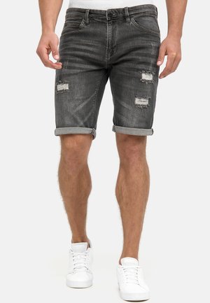 Mann trägt dunkelgraue, abgenutzte, hochgekrempelte Jeansshorts und weiße Turnschuhe, steht mit entspannten Händen an den Seiten.