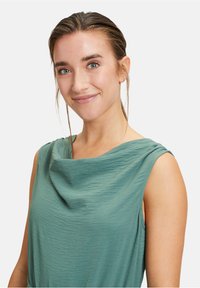 Ärmelloses Oberteil mit Kapuzenkragen aus strukturiertem grünen Stoff, mit drapierter Gestaltung und glatter Oberfläche, das den Schulter- und Halsbereich betont.