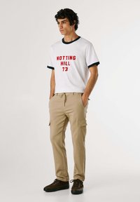 Vit t-shirt med röd text "NOTTING HILL 73", marinblå ringer. Beige cargobyxor med sidofickor. Bruna skor med mörka sulor.