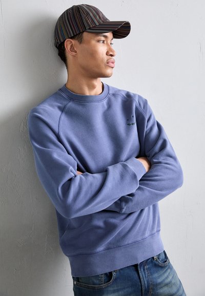 Lyseblå hettegenser med ribbestrikkede mansjetter og midje, med en liten grønn logo. En flerfarget stripete caps er på hodet. Løse denimjeans.