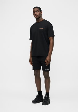 Un homme debout porte un T-shirt noir à manches courtes et un short avec le texte "Les Benjamins", des chaussettes noires et des baskets noires sur fond blanc.