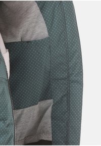 Zelená potištěná látka s malými kruhy, která má měkkou šedou podšívku a hladký zip po okraji. Jsou viditelné více kapsy.