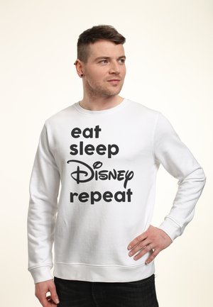 Mężczyzna z krótkimi brązowymi włosami, ubrany w białą bluzę z czarnym napisem „eat sleep Disney repeat”, stoi z jedną ręką na biodrze, patrząc w prawo.