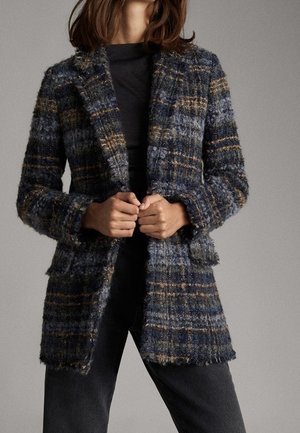 Person wearing a textured plaid blazer in blue, brown, and gray tones over a black shirt and dark pants, hands clasping the blazer front.