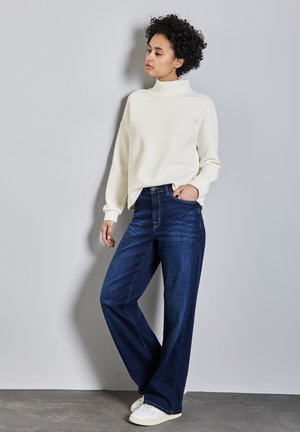 Maglione bianco con collo alto e texture, abbinato a jeans wide-leg in denim scuro a vita alta e sneakers bianche. Lo sfondo è grigio uniforme.